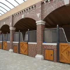 Rangée de stalles de chevaux en briques avec portes en bois et toiture transparente.