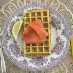 Gaufres salées garnies de saumon fumé et d'un quartier de citron, sur une assiette ornée, avec une fourchette et un couteau à manche jaune.