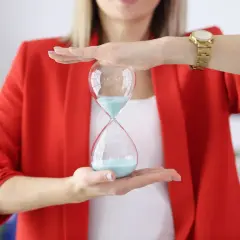 Kobieta w czerwonej marynarce trzyma klepsydrę z niebieskim piaskiem. Czas płynie, a figura klepsydra symbolizuje jego upływ.