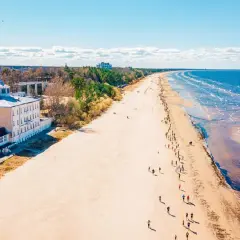 Szeroka, piaszczysta plaża na Łotwie, z budynkami przy brzegu i ludźmi spacerującymi wzdłuż morza.