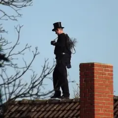 Ein Schornsteinfeger in Zylinder und schwarzer Kleidung steht auf einem Dach. Er trägt seine Bürste und Kette bei sich. Man fragt sich, wie oft kommt der Schornsteinfeger wohl?