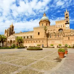 La maestosa cattedrale di Palermo si erge sotto un cielo azzurro con nuvole bianche, con la sua architettura imponente e il suo cortile lastricato.