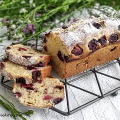 Szybkie ciasto z czereśniami, posypane cukrem pudrem, prezentuje się apetycznie na metalowej podstawce. W tle kwitną polne kwiaty.