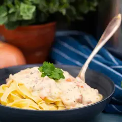En skål med tagliatelle täckt av krämig ost och skinksås, toppad med persilja. En gaffel sticker upp ur pastan.
