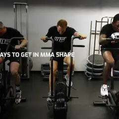 Three men with impressive fighters physique train on air bikes in a gym, showcasing their dedication to peak physical condition.