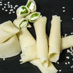 Rotolini di crepes con farina di riso, ripieni di formaggio fresco e basilico, sparsi su un tavolo scuro con semi di sesamo.
