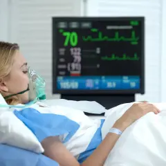 Kobieta w masce tlenowej leży w szpitalnym łóżku, monitor pokazuje parametry życiowe. Jej stan budzi niepokój, być może to rzucawka.