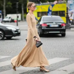 Elegancka, beżowa sukienka maxi, idealna na miejskie stylizacje. Kobieta w okularach przeciwsłonecznych i małej czarnej torebce przechodzi przez pasy.