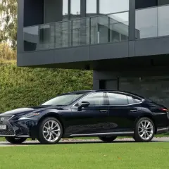 Elegancki, ciemnogranatowy Lexus LS 500 zaparkowany przed nowoczesnym domem z dużymi oknami i zielonym ogrodem.