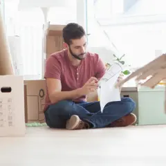 Hombre revisa un plano rodeado de cajas, preparándose para la mudanza. Podría ser el **contrato compra venta** de su nuevo hogar.