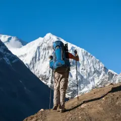 Un excursionista con mochila azul asciende un sendero polvoriento, bastones en mano, ante majestuosas montañas nevadas. Una aventura de trekking Manaslu.