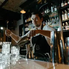 Barman przygotowuje drinka, zastanawiając się, ile zarabia dobry barman. Za nim półki z alkoholem.