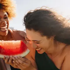 Zwei lachende Frauen teilen ein Stück Wassermelone. Perfekt für einen heißen Tag, da Wassermelone kalorienarm ist und erfrischt.
