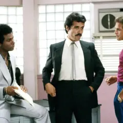 Trzech mężczyzn, w tym policjanci z Miami obsada, w tym Sonny Crockett i Ricardo Tubbs, rozmawiają w biurze.
