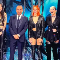 Micky Paiano con un completo scuro, affiancato da quattro donne con outfit in pelle audaci e colorati, sorridono sul palco.