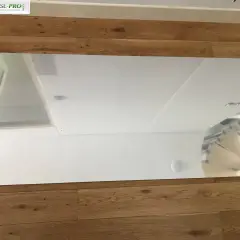 Un grand panneau de dibond miroir reflète le plafond et un mur blanc, posé sur un parquet en bois.