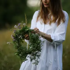 Kobieta w białej sukni trzyma wianek z polnych kwiatów na tle zielonej łąki.