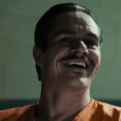 Lalo Salamanca ler skrattande i en orange fängelsedräkt, hans mustasch framträder tydligt.