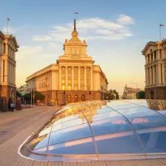 Zachód słońca nad placem w Sofii w Bułgarii. Monumentalne budynki i szklana konstrukcja na pierwszym planie.