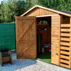 Duży domek narzędziowy z otwartymi drzwiami, w środku kosiarka i narzędzia. Obok stos drewna.