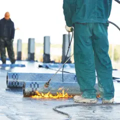 Arbeiter schweißen Bitumenbahnen, um ein Flachdach abzudichten. Flammen erhitzen das Material für eine sichere Verbindung.