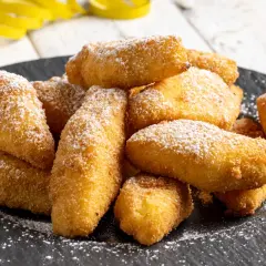 Un mucchio di dorate crema fritta, cosparse di zucchero a velo, su un piatto scuro.