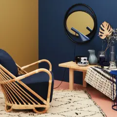 Ein Rattan-Sessel mit dunkelblauem Kissen steht auf einem flauschigen Teppich. Welche Farbe passt zu Gold? Hier: ein Spiegel mit goldenem Rahmen und ein gelber Wandbereich.