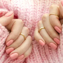 Dłonie z różowym manicure i złotymi pierścionkami na tle różowego swetra.
