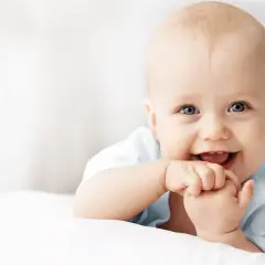 Uśmiechnięte niemowlę z niebieskimi oczami, które właśnie przeżywa swój pierwszy skok rozwojowy, z radością wkłada rączki do buzi.