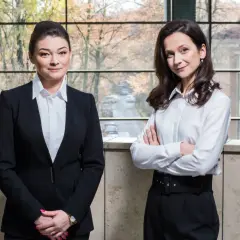 Dwie kobiety w strojach biznesowych stoją przed oknem z widokiem na miasto.