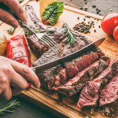 Une main coupe un succulent côte de bœuf barbecue sur une planche en bois, accompagné de pommes de terre rôties et de poivrons grillés.