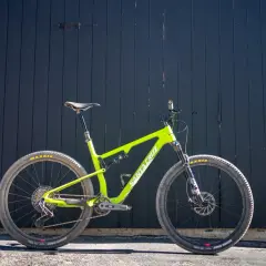 A bright green Santa Cruz Blur mountain bike is ready for a trail review, leaning against a dark wooden wall.