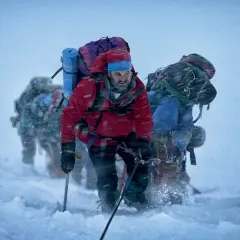 Grupa himalaistów w czerwonych i niebieskich kurtkach, z plecakami, wspina się w śnieżycy. Scena jak z filmów o himalaistach.