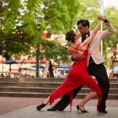 Coppia in abito elegante e vestito rosso esegue una sensuale danza latina all'aperto, tra alberi e gradinate.