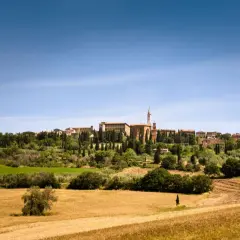 Un borgo medievale domina la Val d'Orcia, circondato da campi dorati e cipressi sotto un cielo azzurro.