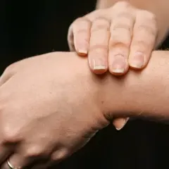 Dłoń z widocznym, twardym guzem na wewnętrznej stronie nadgarstka. Palce drugiej dłoni delikatnie dotykają tego miejsca.