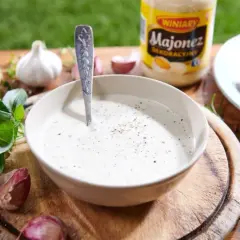 Miseczka z gęstym, białym sosem, łyżka, czosnek, zioła i słoik majonezu.
