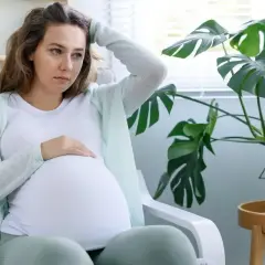 Zmartwiona ciężarna siedzi w fotelu, dotykając brzucha. W tle duża monstera. Podwyższone obawy o przyszłość.