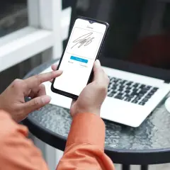 Persona usando su móvil para firmar online documento. En la mesa hay un portátil y una taza de café.