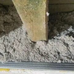 Luźna celuloza w szczelinach, drewniana belka i metalowa siatka. Wady ocieplenia celulozą widoczne przez nierównomierne rozłożenie materiału.