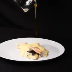 Ein Koch übergießt Fisch mit Spargel und Kaviar mit einer cremigen Sauce. Die Sauce sieht aus, als wäre die Sahne geronnen, was dem Gericht eine interessante Textur verleiht.