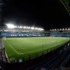 Piłkarski stadion Molde nocą, oświetlony jaskrawymi światłami, z pustymi niebieskimi trybunami i zieloną murawą.