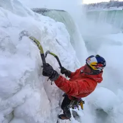 Wspinaczka lodowa przy zamarzniętych wodospadach. Człowiek w pomarańczowej kurtce i kasku z logo Red Bull używa czekana, by pokonać lodową ścianę.