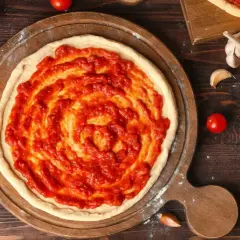 Une pâte à pizza crue est étalée sur une planche en bois, recouverte d'une sauce tomate maison. La question quelle sauce tomate pour pizza est au cœur de cette préparation.