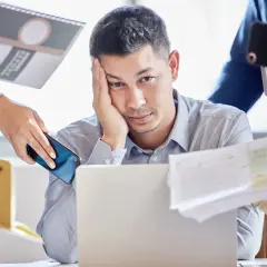 Zmęczony mężczyzna przy laptopie, otoczony dokumentami i telefonami. Czy to właśnie multitasking co to znaczy?