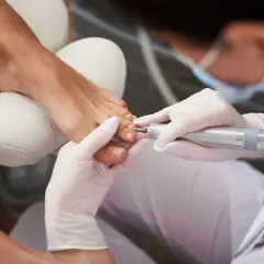 Profesjonalny pedicure frezarkowy w trakcie. Dłoń w białej rękawiczce używa frezarki do pielęgnacji paznokci stóp.