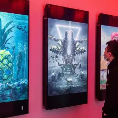 Ein Mann betrachtet digitale Kunstwerke, die an einer Wand hängen. Die Werke zeigen surreale Szenen, wie eine Ananas unter Wasser mit Skeletten und eine symmetrische Figur mit vielen Armen. Diese Werke stammen von einem berühmten Grafik Künstler.