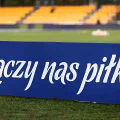 Niebieska tablica z napisem "łączy nas piłka" na tle stadionu. To motto, które jednoczy kibiców i prezesa PZPN.