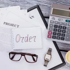 Na biurku leżą dokumenty, notatnik z napisem "Order", kalkulator, okulary, zegarek i kawa. To wszystko, co potrzebne do pracy, gdy zastanawiasz się, moq co to.