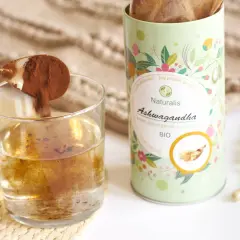 Łyżeczka z proszkiem ashwagandhy wsypywana do szklanki wody. Opakowanie Naturalis Ashwagandha BIO obok.
